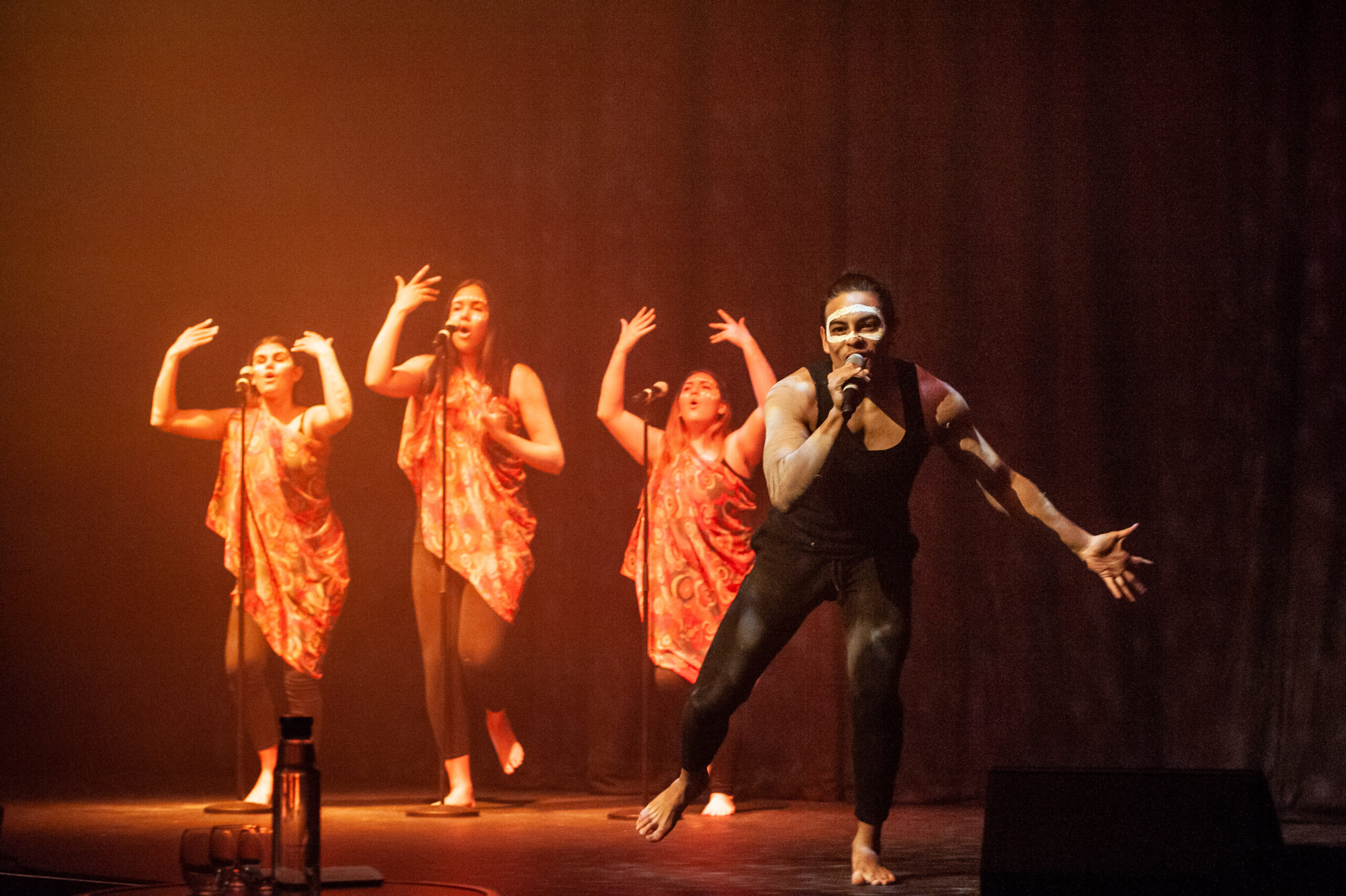 Four performers are on stage. Three are standing in the background singing, wearing similar garments, while one performer in the foreground is singing and reaching towards the audience.