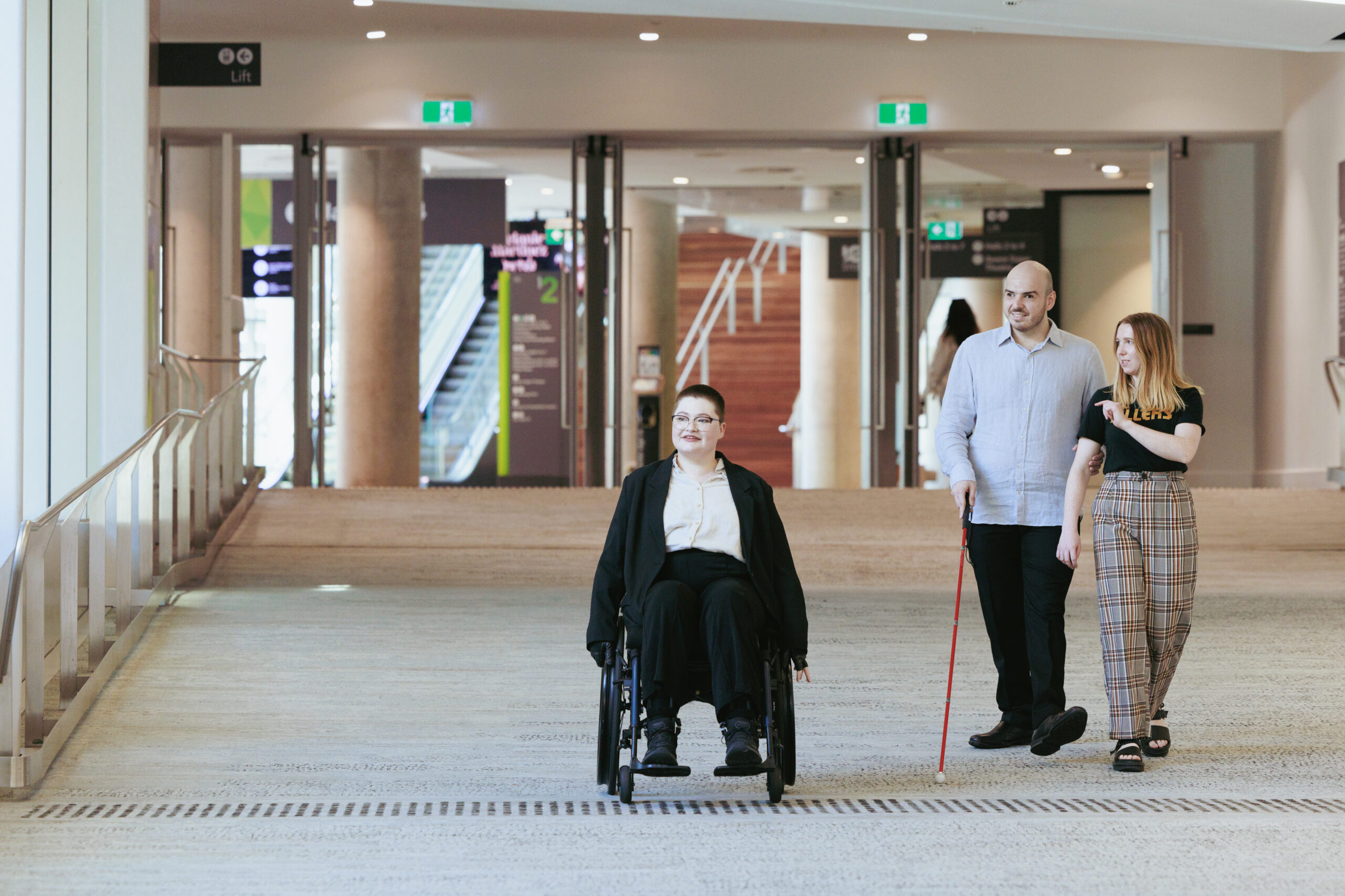 A person in a wheelchair, a person using a white cane, and a person walking together in a well-lit indoor public space.